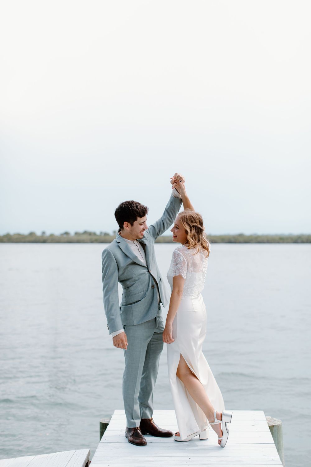 A couple dances together on a dock overlooking calm waters, wearing formal wedding attire.