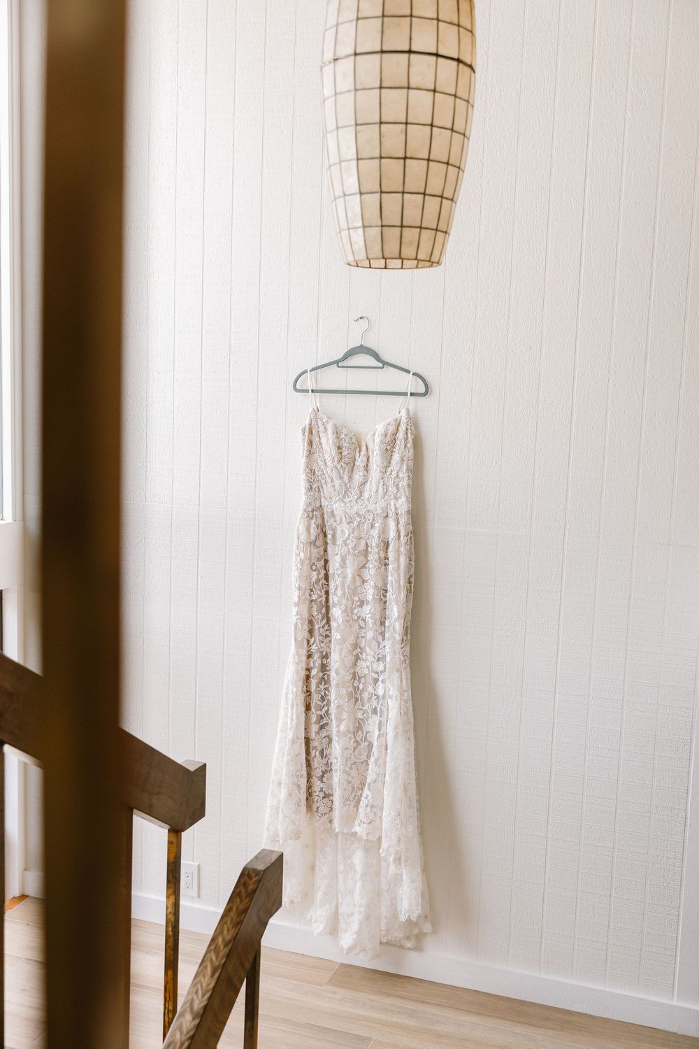 A delicate lace wedding dress hangs on display under a modern pendant light.