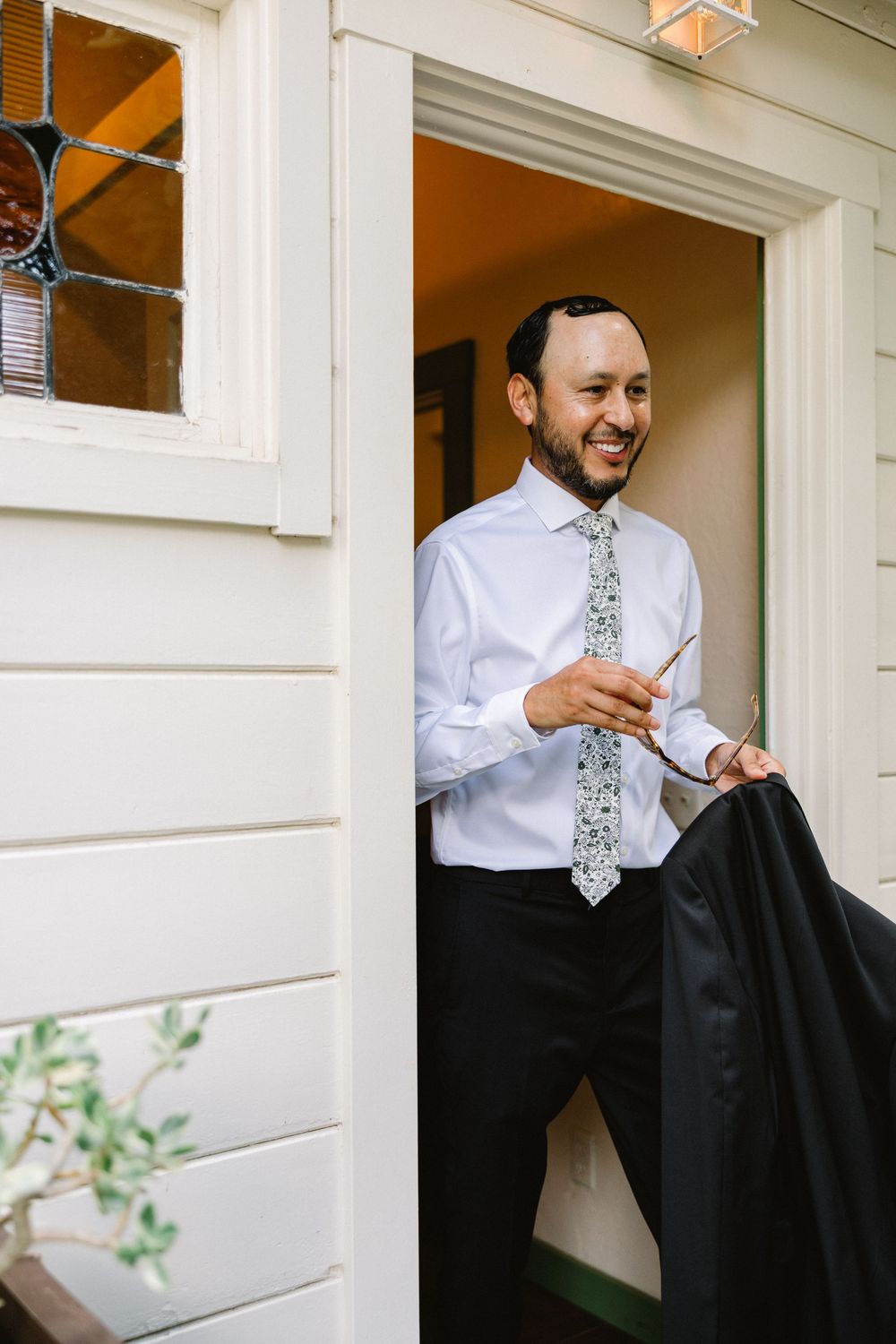 A smiling wedding guest in formal attire holding a suit jacket in a doorway.