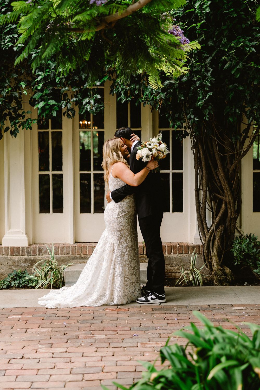 A newlywed couple embraces outside a white columned building with climbing vines.