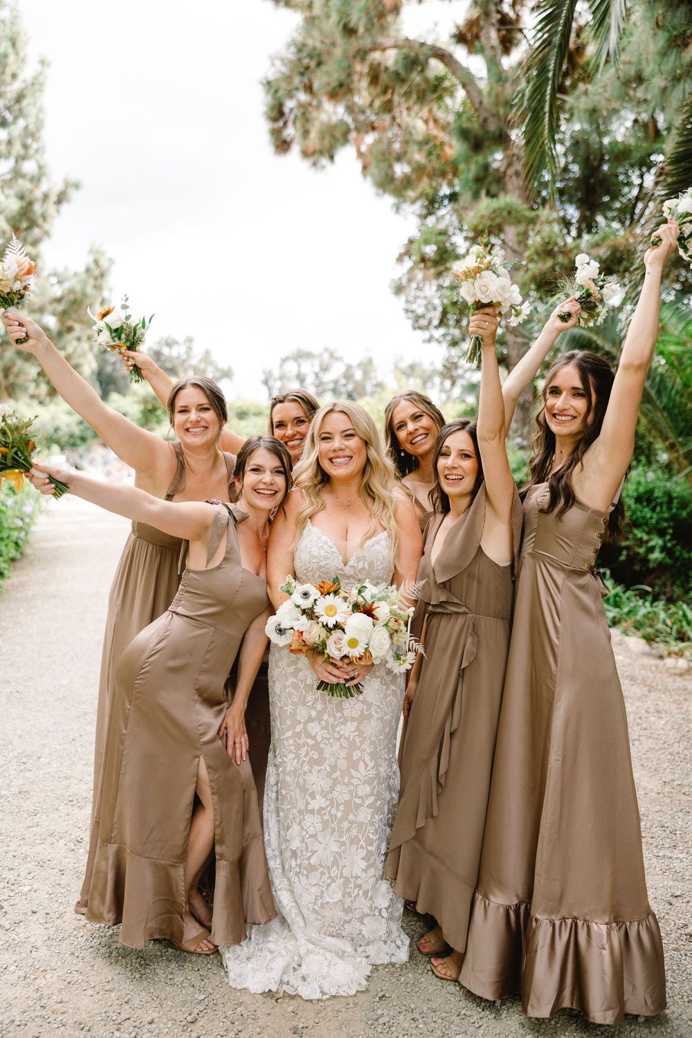 Wedding party poses outdoors with bridesmaids in tan dresses celebrating with raised arms.