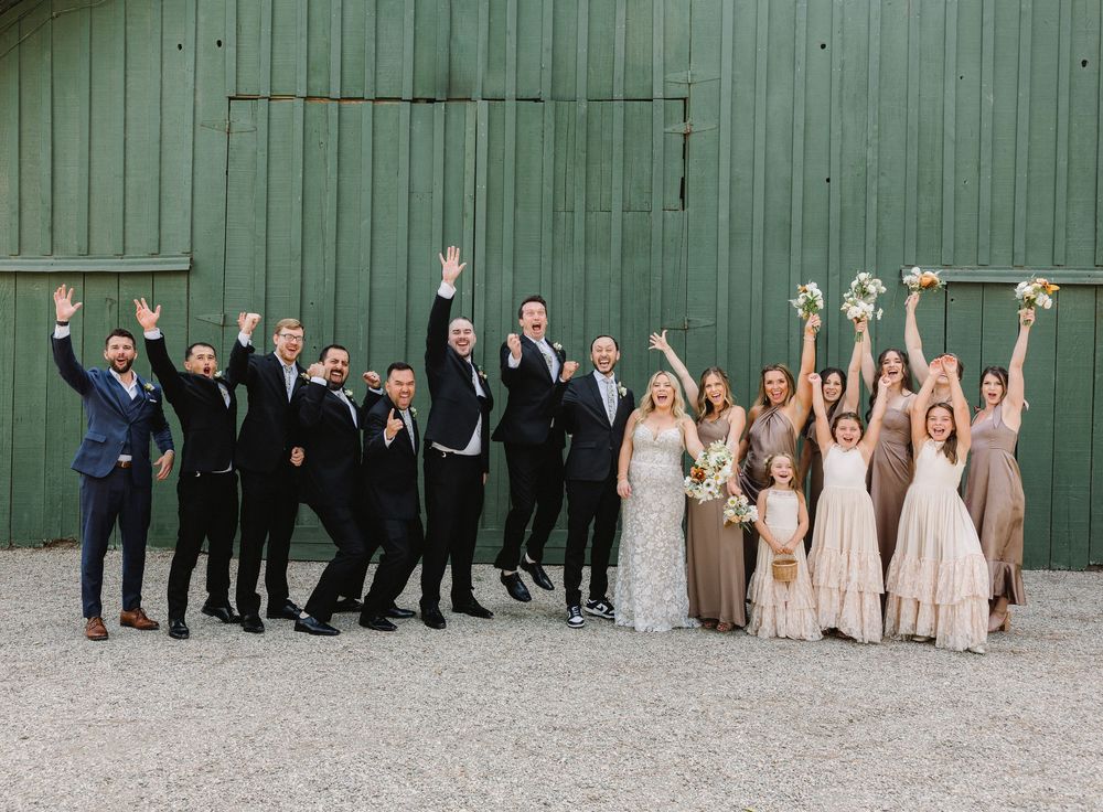 Wedding party poses against a green barn with everyone raising their arms in celebration.