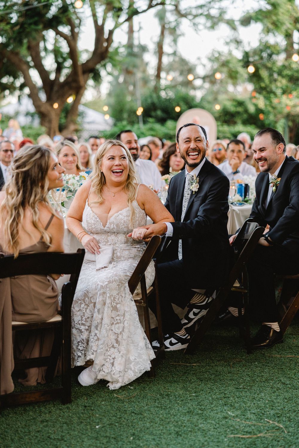 Wedding guests laugh during an outdoor reception under string lights and trees.