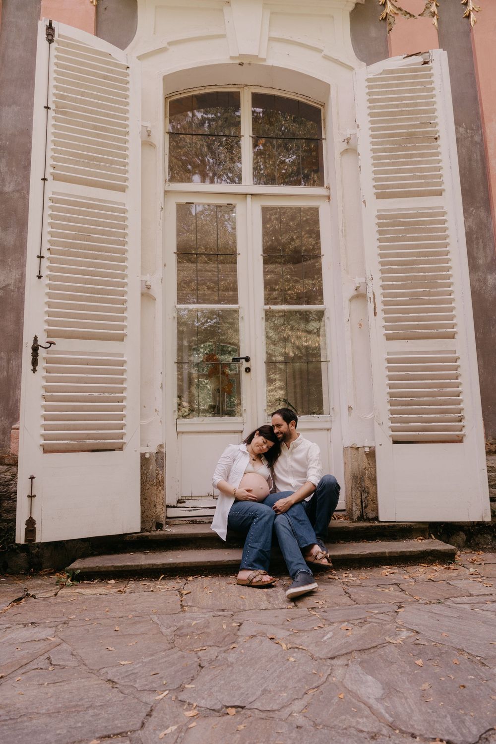 Couple sitting on steps in front of vintage white shuttered door, sharing a tender moment during maternity photo session.
