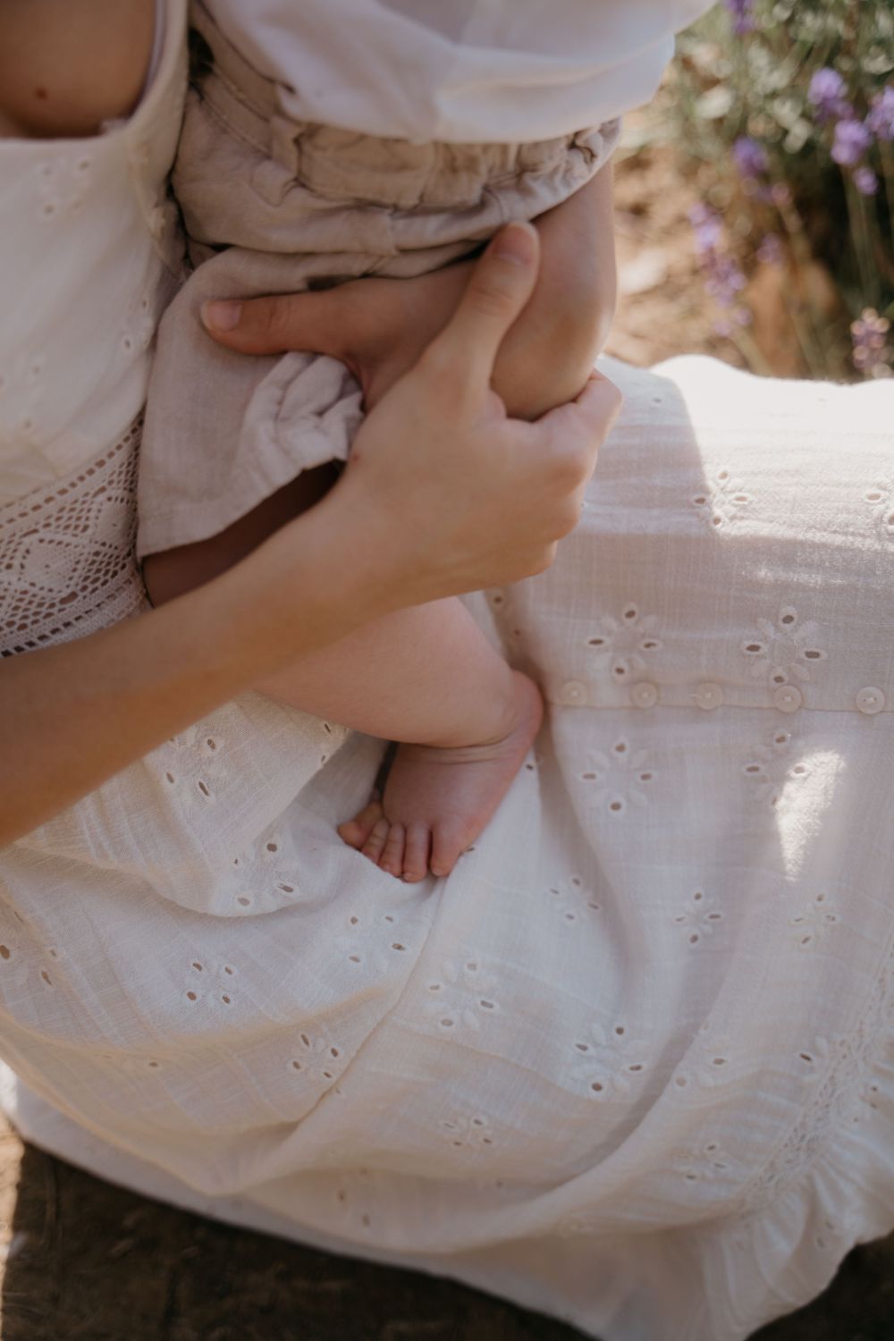 A mother in a white eyelet dress tenderly holds her newborn baby's tiny feet outdoors near blooming flowers.