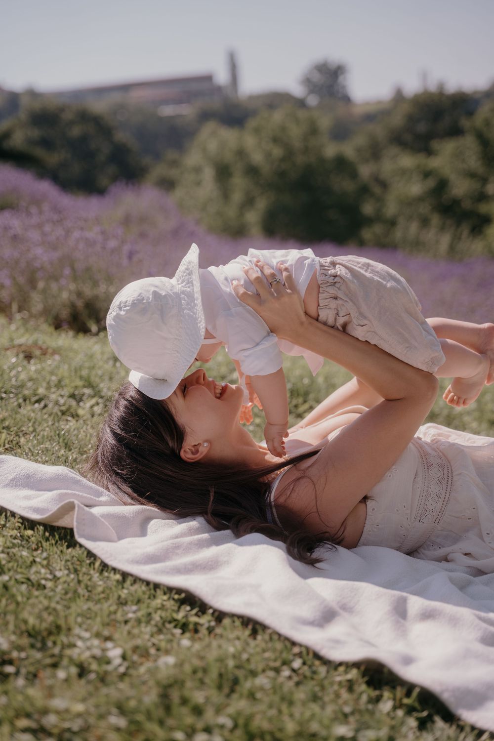 A mother lies on a blanket in a lavender field, joyfully lifting her baby in white outfit up above her face.