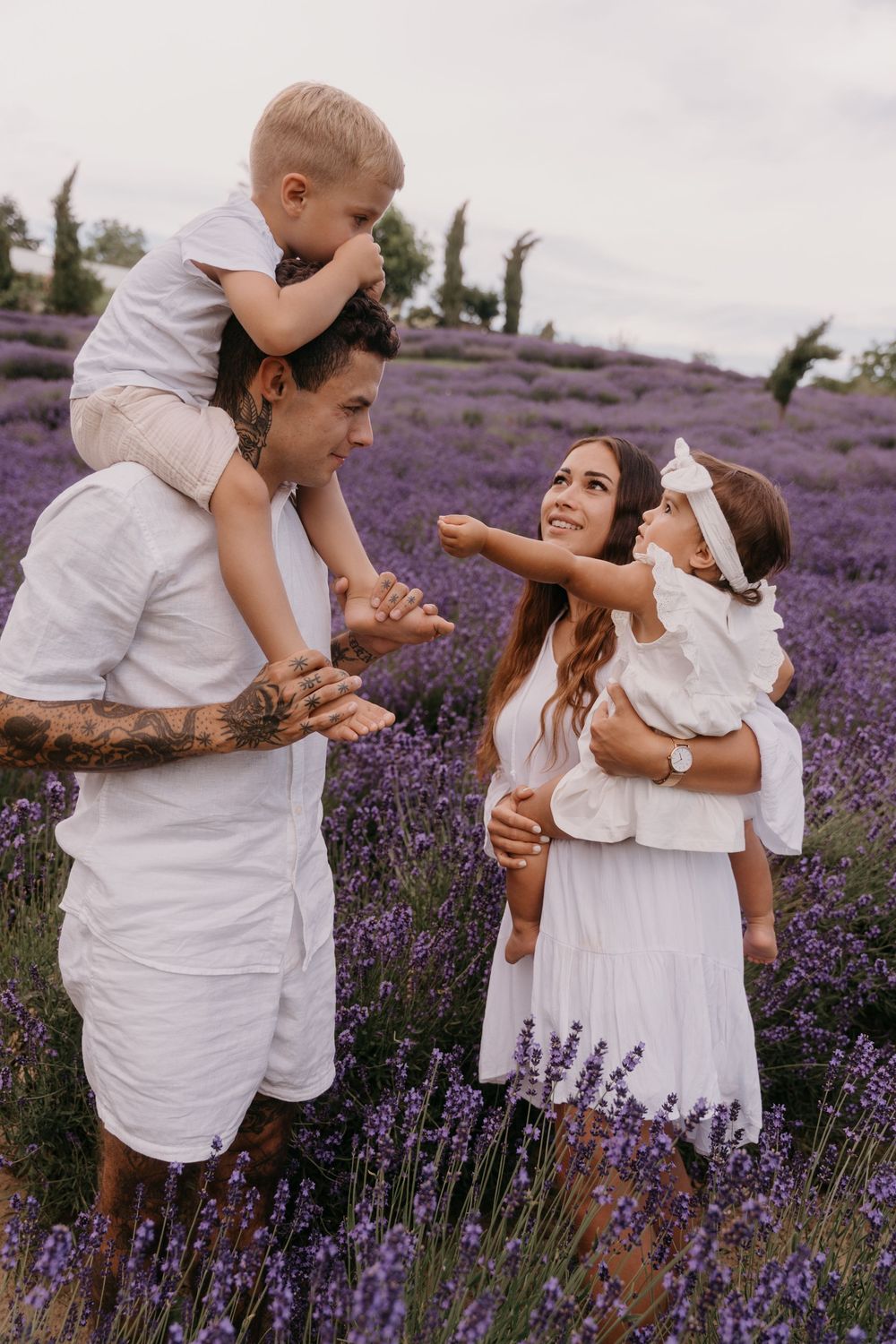 Family in white outfits enjoying a sunny day in a blooming lavender field, with children laughing and reaching out.
