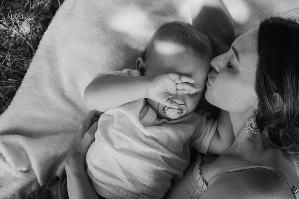 Black and white photo of a mother kissing her baby lying on a blanket outdoors in soft natural light.