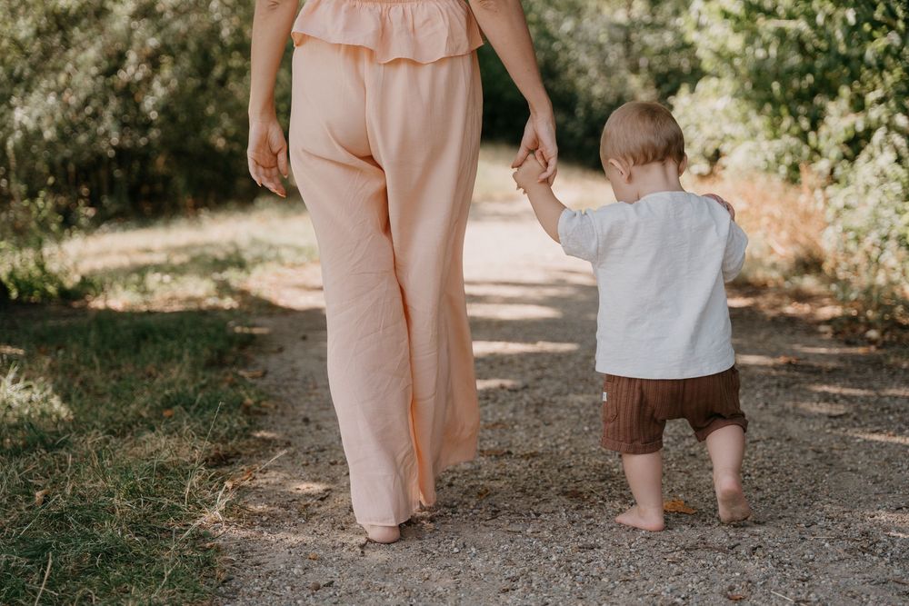 A mother in a peach outfit holds hands with her toddler on a dirt path surrounded by green trees.