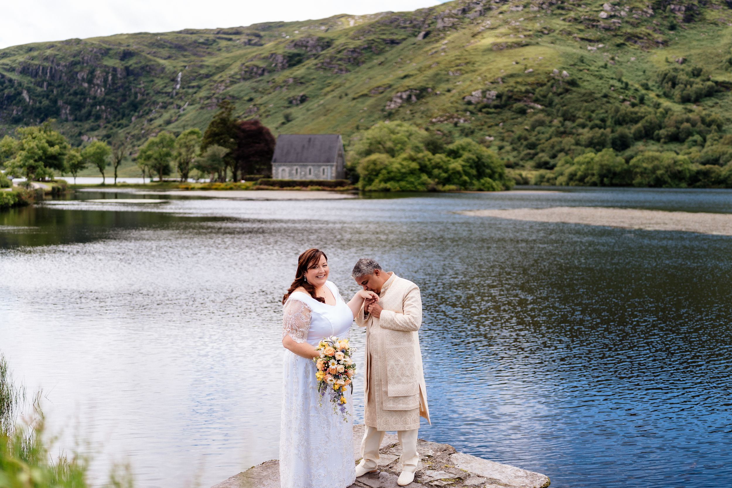 A couple in wedding attire walks hand in hand along a peaceful lake shore with mountains in the background.