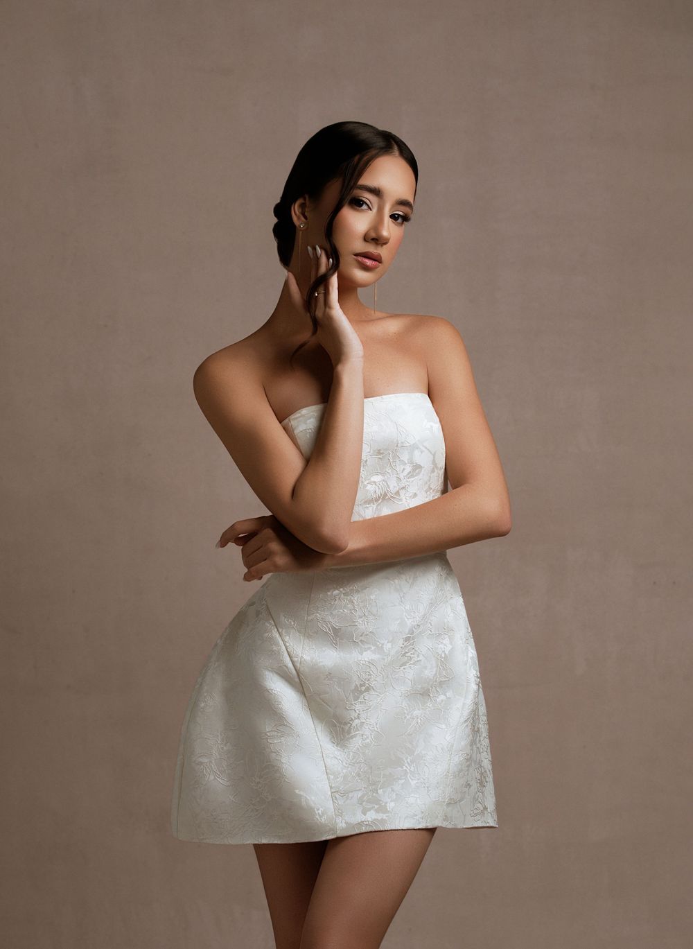 A model in a short white strapless dress poses elegantly against a neutral brown background in a fashion photoshoot.