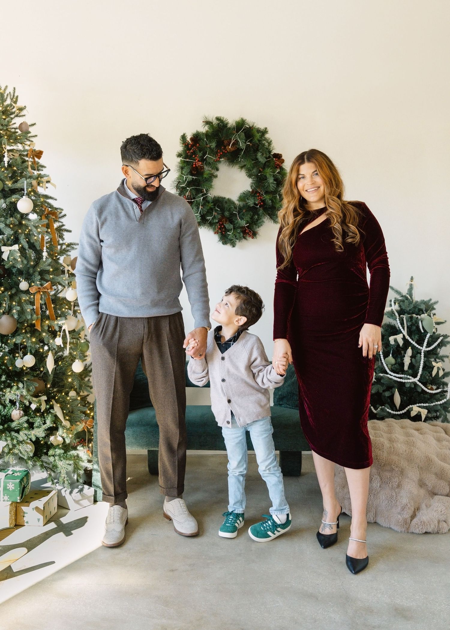 Woman in white outfit sits near Christmas tree watching toddler play on wooden rocking horse.