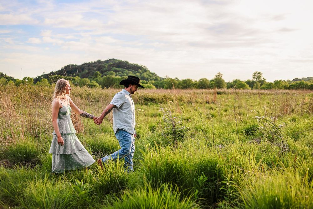 A lifestyle image of a couple walking through a field with hills in the background at golden hour sunset.