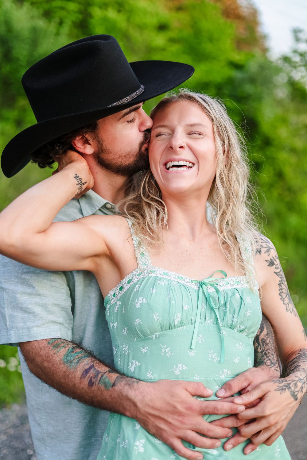 A candid image of a young couple holding each other laughing while he kisses her on the cheek.