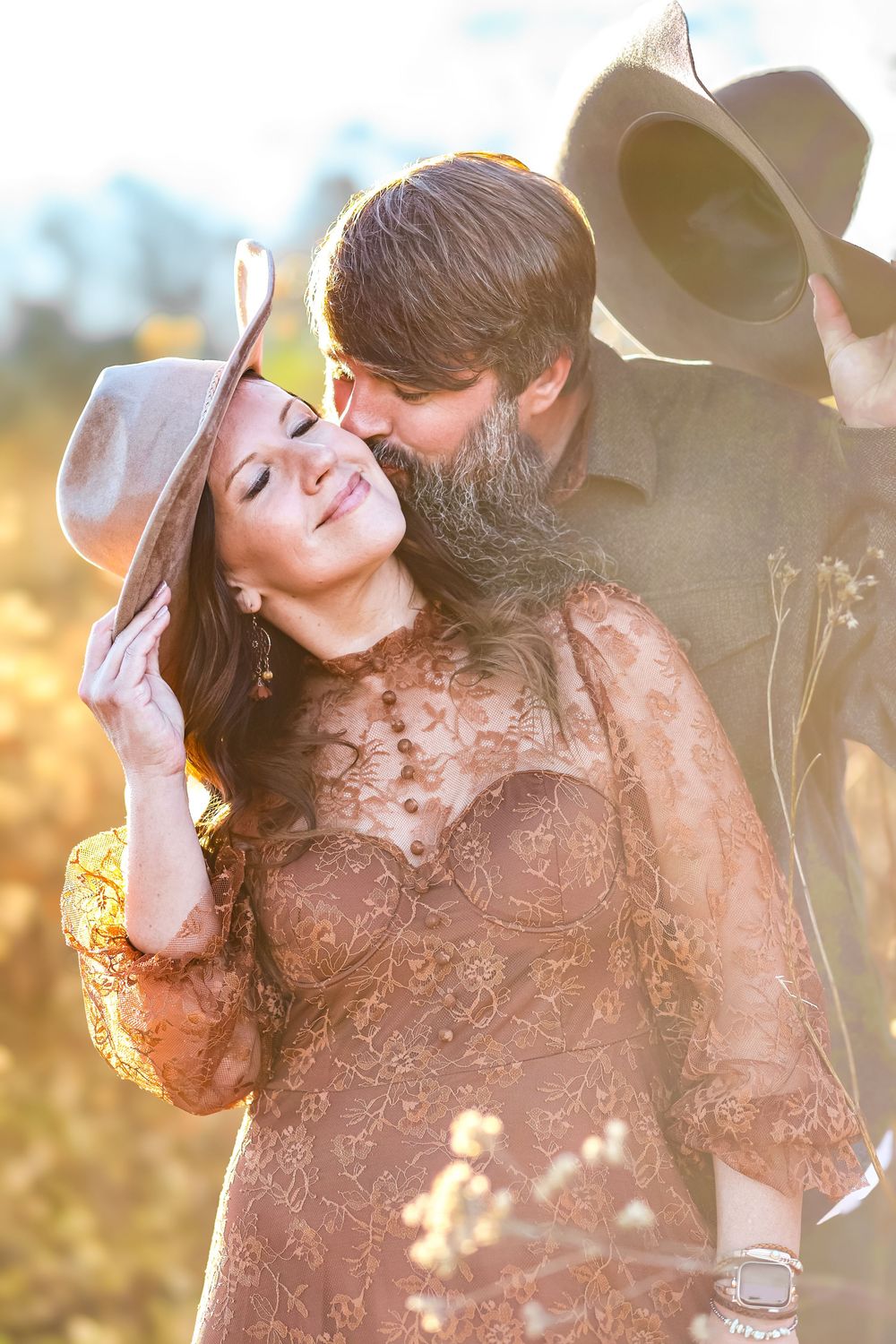 A candid western themed couple wearing cowboy hats kissing his wife on her cheek while she holds her hat in the hazy sunlight smiling.