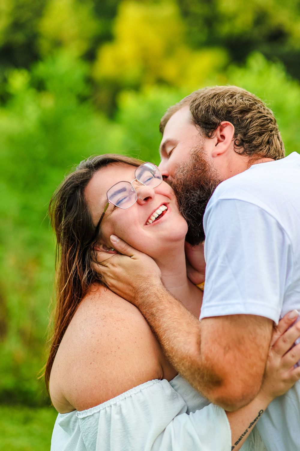 A candid image of a couple in a green field. The man gives a kiss on the side of her face smiling.