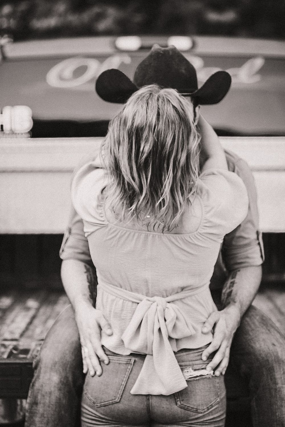 A candid picture of a couple posing on the back of a tailgate facing each other with a cowboy hat on (Black and white)