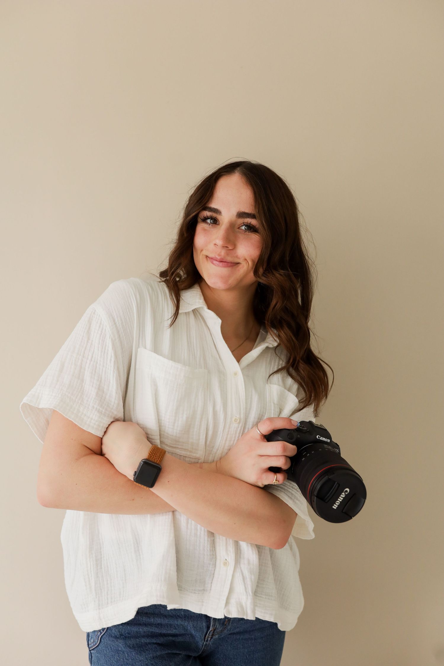 Young female photographer smiles holding a Canon DSLR camera in a white linen shirt.