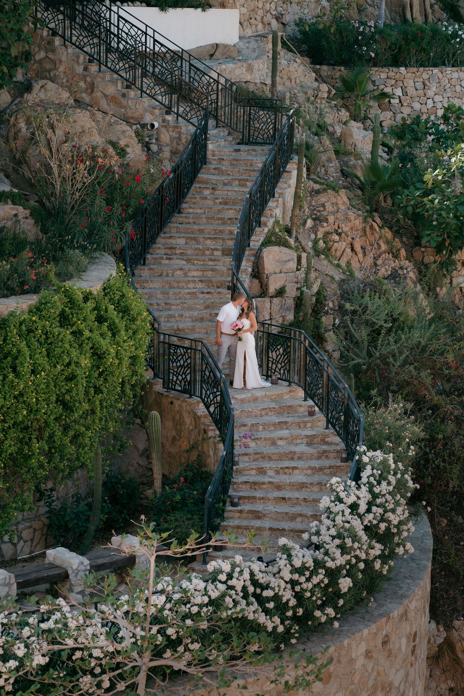 Stone stairway with ornate black iron railing winds up through lush garden with blooming white flowers and greenery.