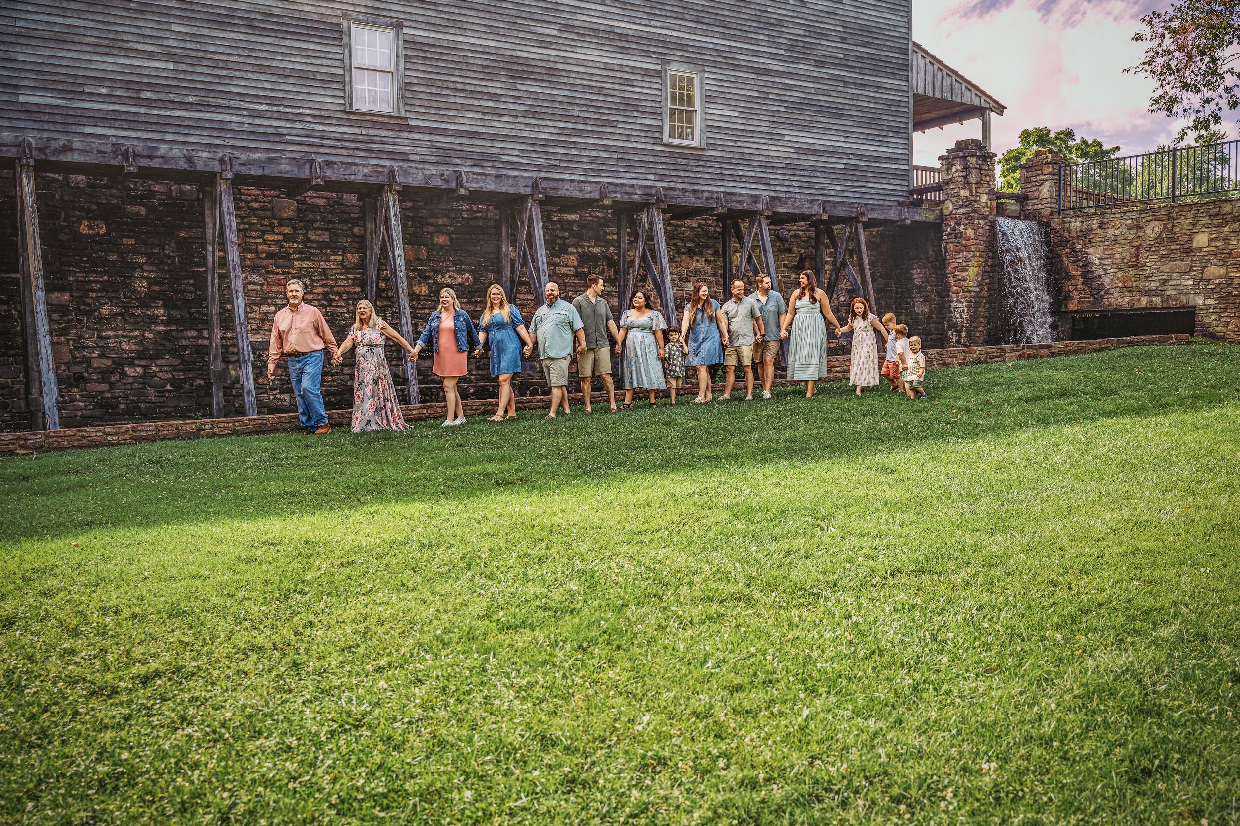 A panoramic view of people standing by a historic wooden building with green lawn in the foreground.