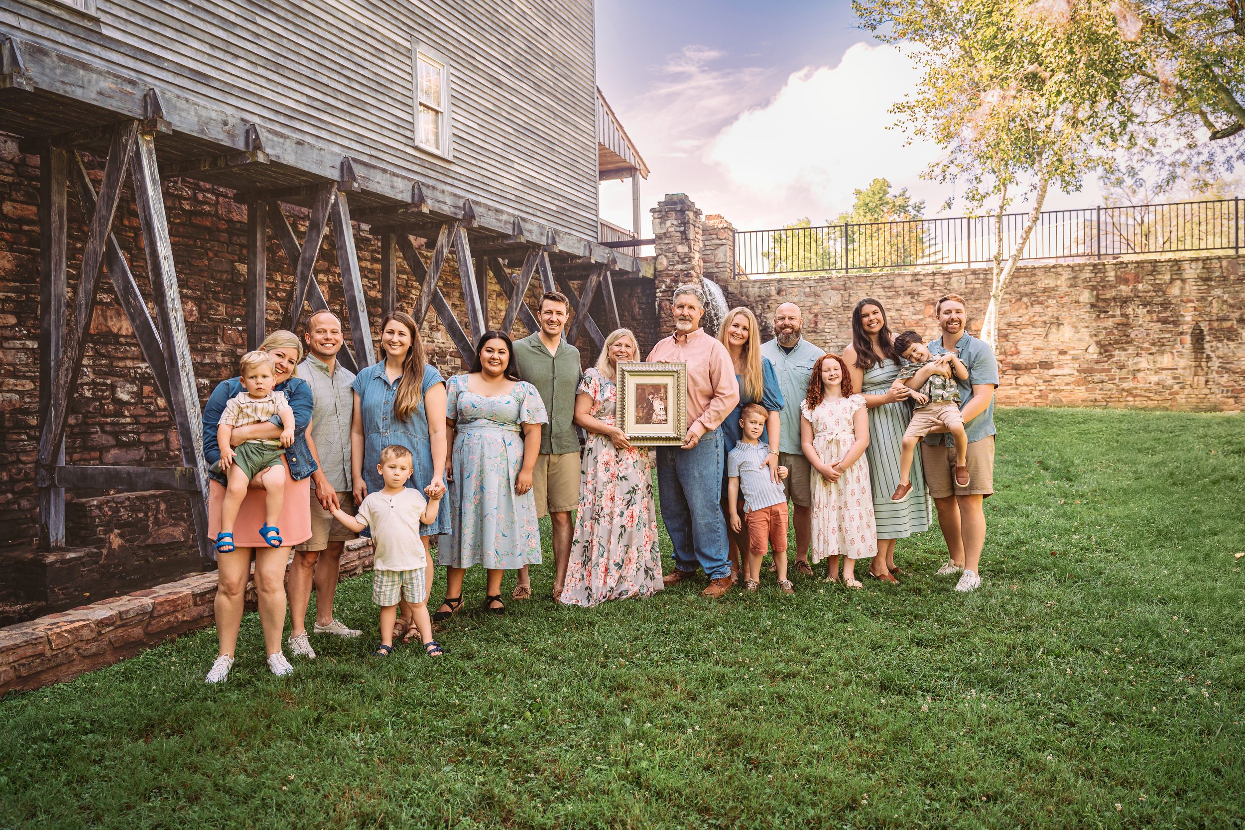A group gathering poses together on a lawn against a rustic brick wall and wooden structure backdrop.