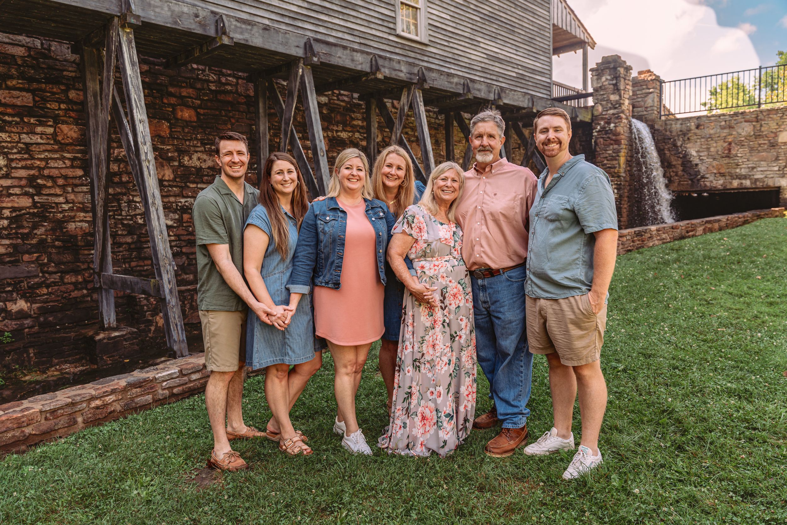 Group of people sitting together on a grassy slope in front of a rustic wooden barn wall.
