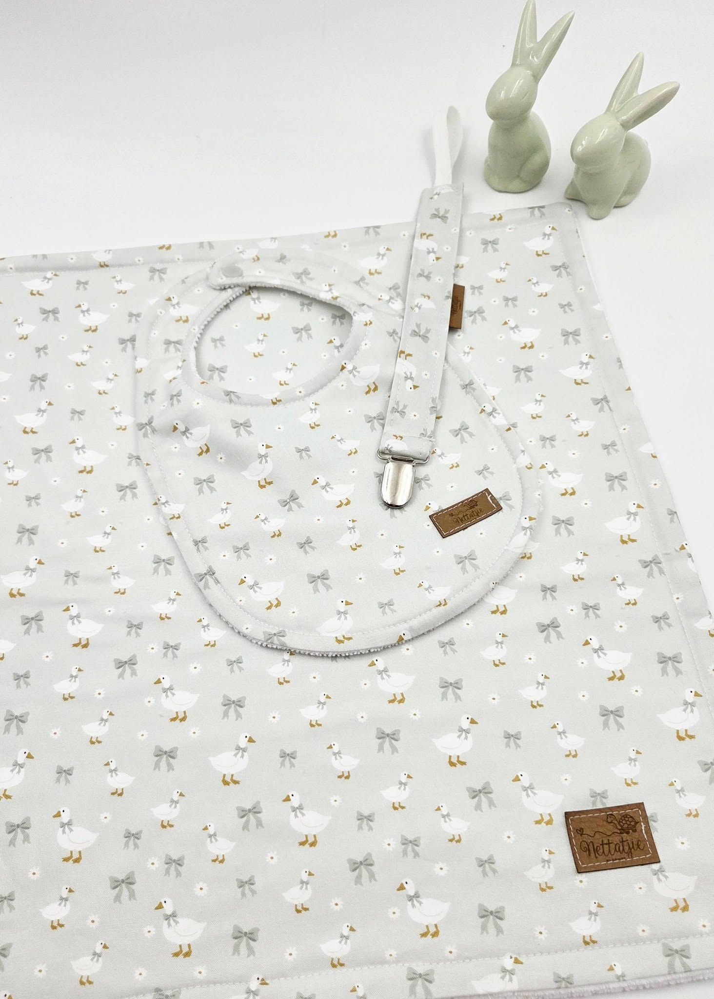 Baby gift set with floral bow print blanket, bib, and pacifier clip, displayed with ceramic bunny figurines.