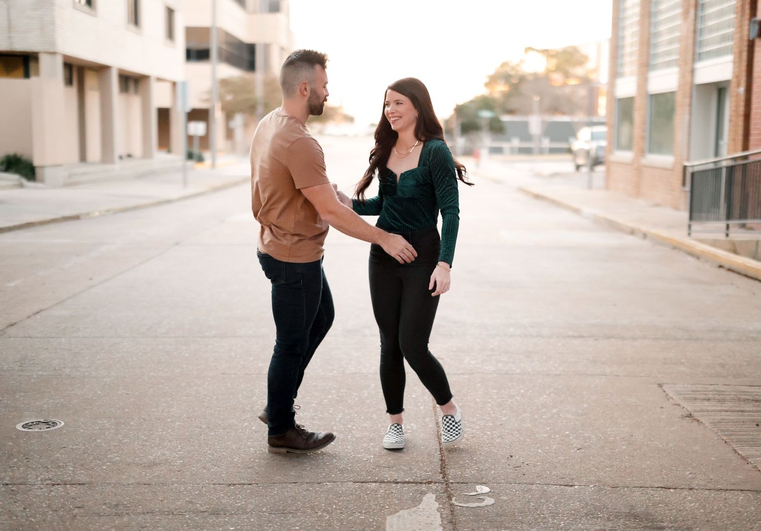 Two people walking and talking together on a downtown sidewalk at sunset, one wearing a brown jacket and one in green.