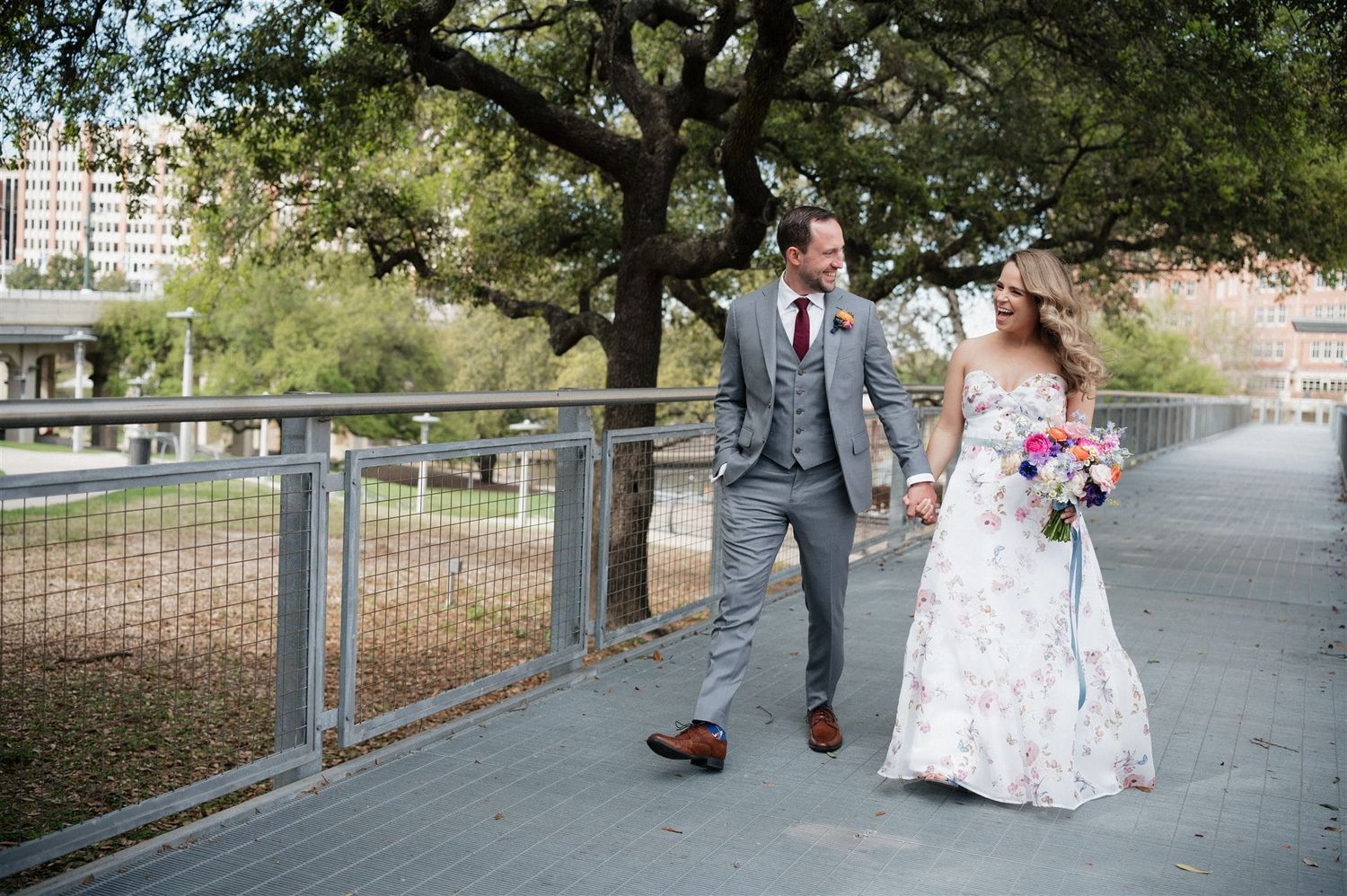 A couple walks along a waterfront path holding hands on their wedding day, with trees and buildings in the background.