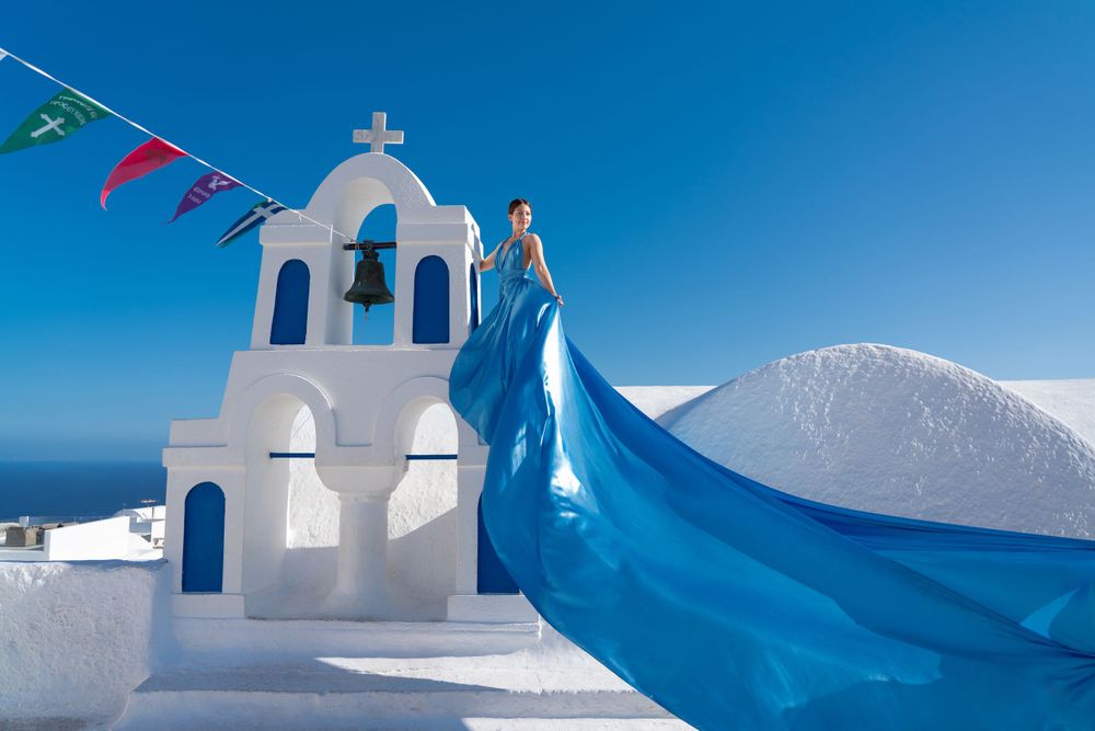 Dramatic blue dress billows against white church bell tower and Mediterranean sky.