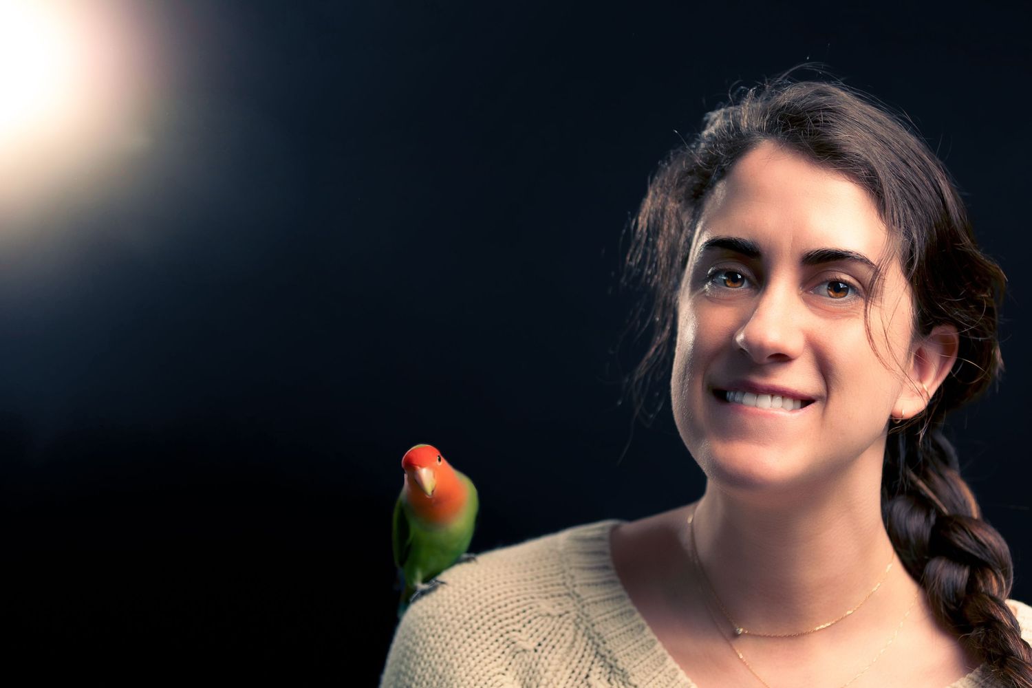 A small orange and green lovebird perches near someone in a beige sweater against a dark background with lens flare.