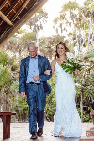 Wedding couple walking under a thatched roof structure surrounded by palm trees, Papaya Playa Tulum Wedding.