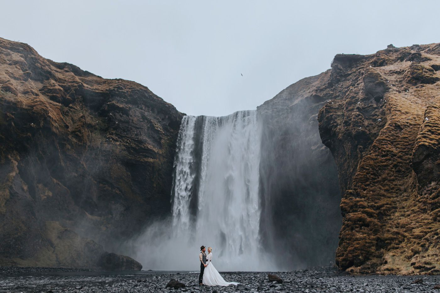 A stunningly small silhouette of newlyweds against a massive Icelandic waterfall.
