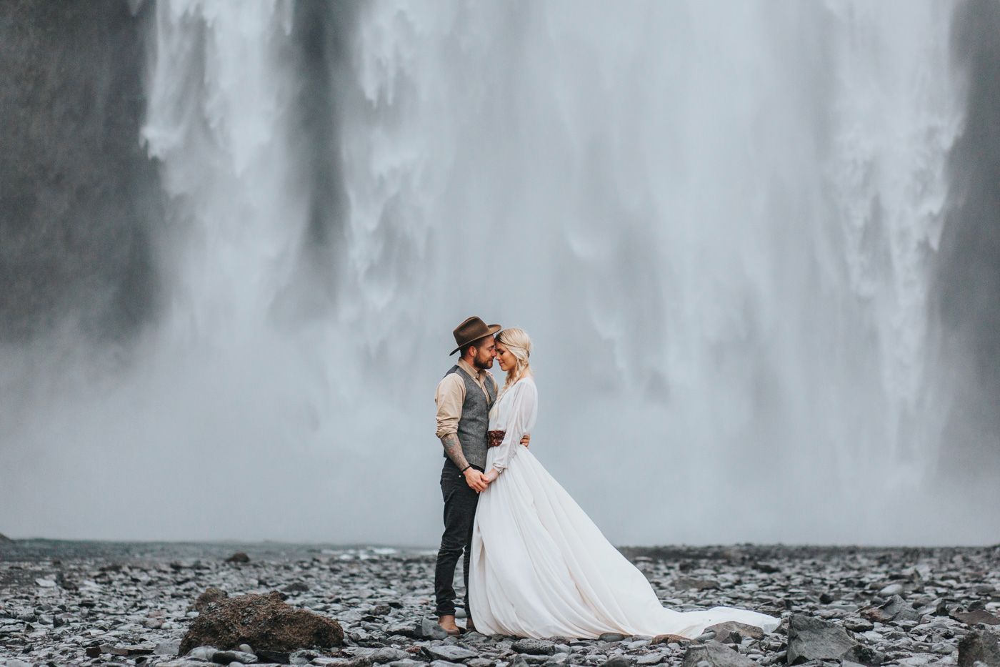 A wedding couple stands on rocky ground before a towering misty waterfall in Iceland.