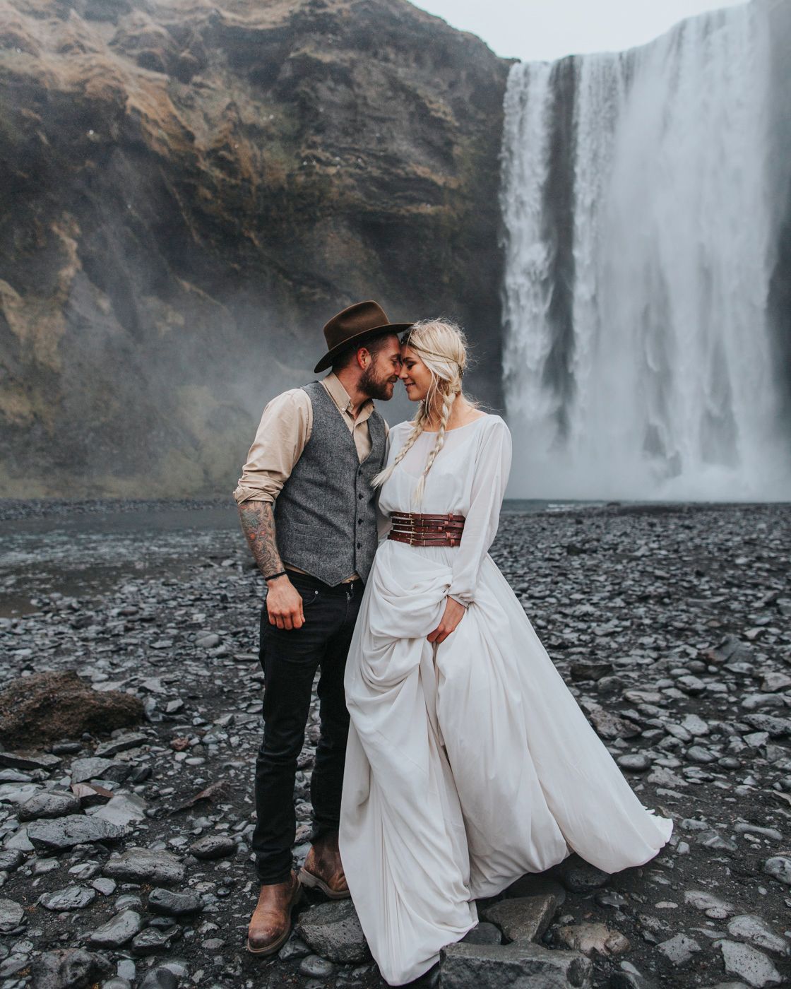Romantic wedding couple embraces in front of majestic waterfall on rocky beach in Iceland.