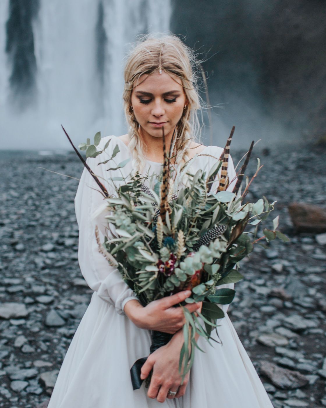 Ethereal bridal portrait with natural bouquet against moody waterfall landscape.