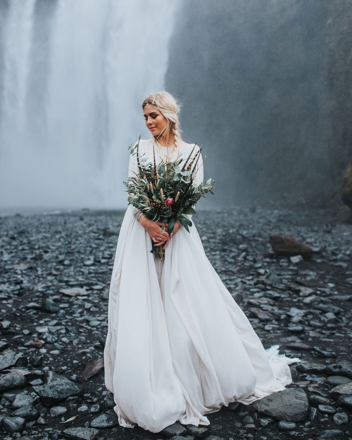 Bride holds rustic bouquet while standing on rocky shore near misty waterfall.