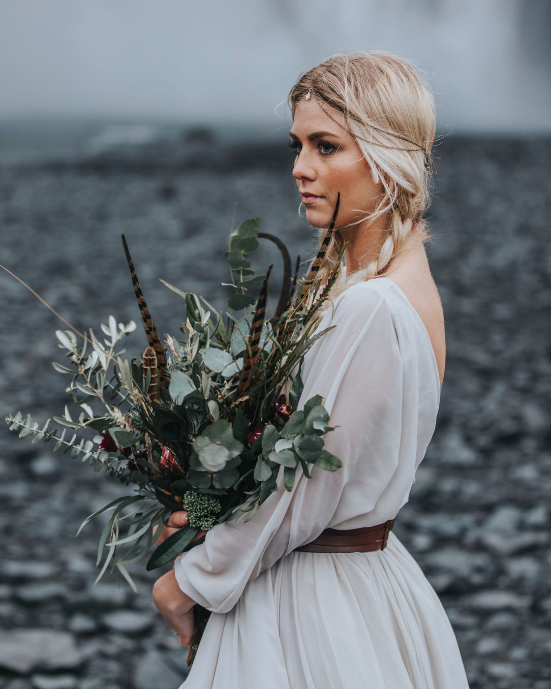 Side profile of bride with rustic bouquet wearing belted white dress near waterfall.