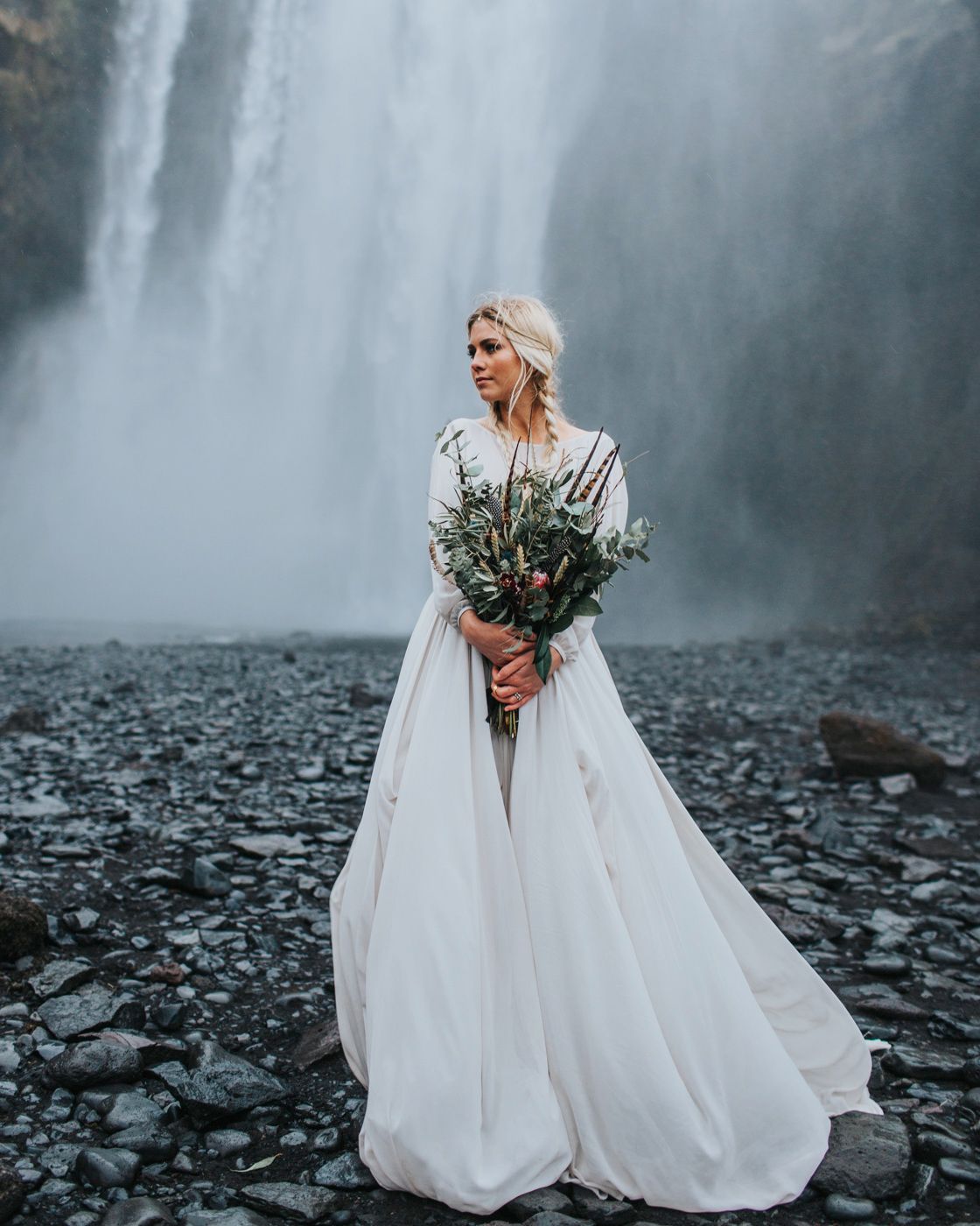 Elegant bride in flowing dress holds wild bouquet against dramatic waterfall setting.
