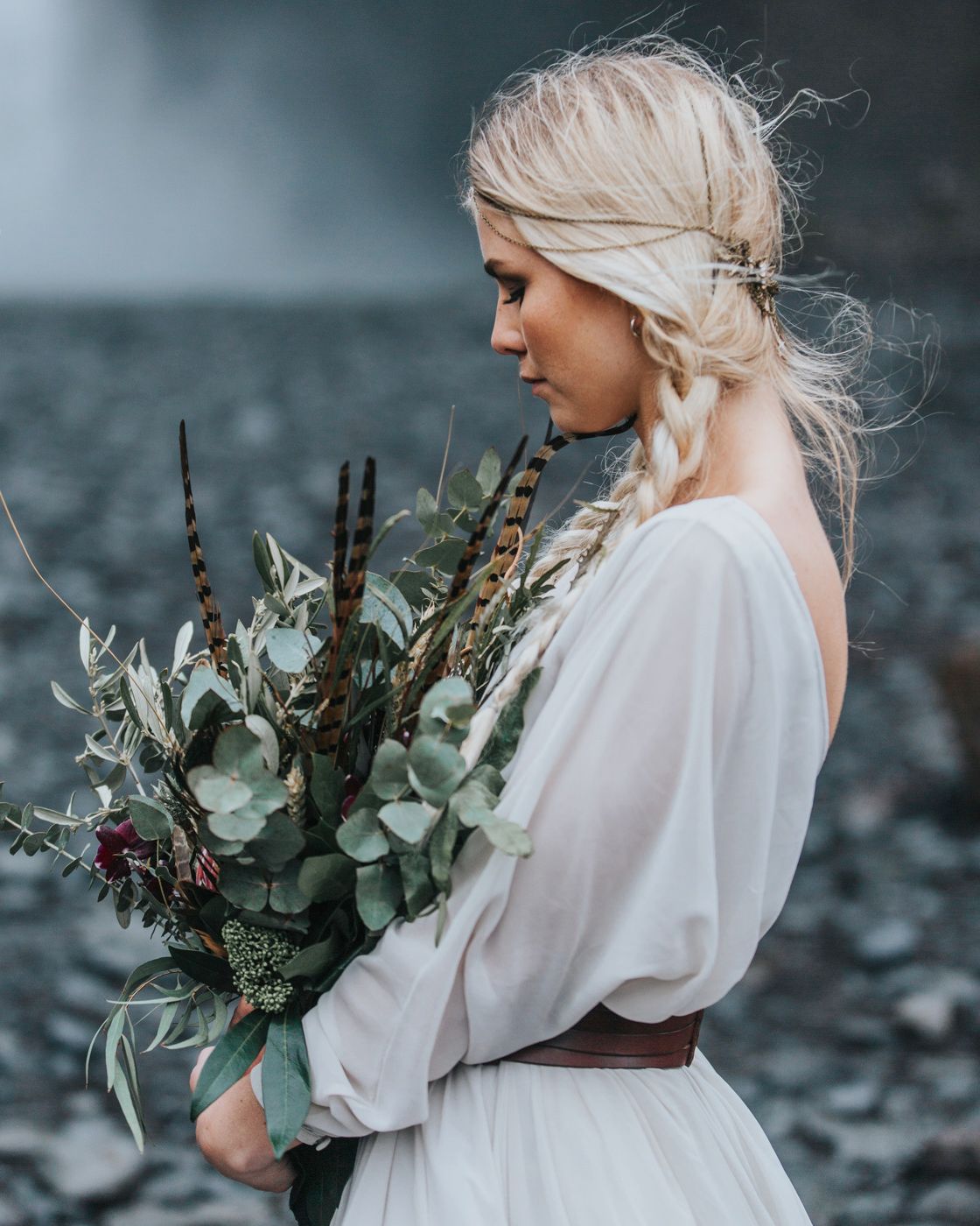Dreamy side view of bride holding eucalyptus bouquet near misty waterfall.