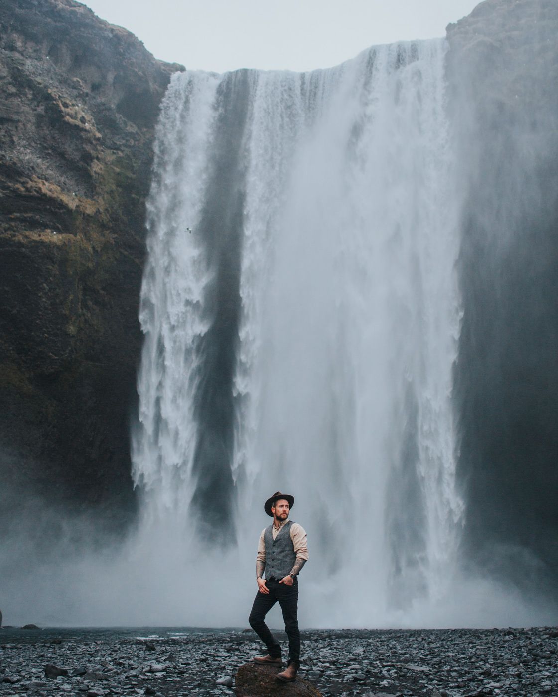 Lone figure in hat and vest poses dramatically before towering waterfall.