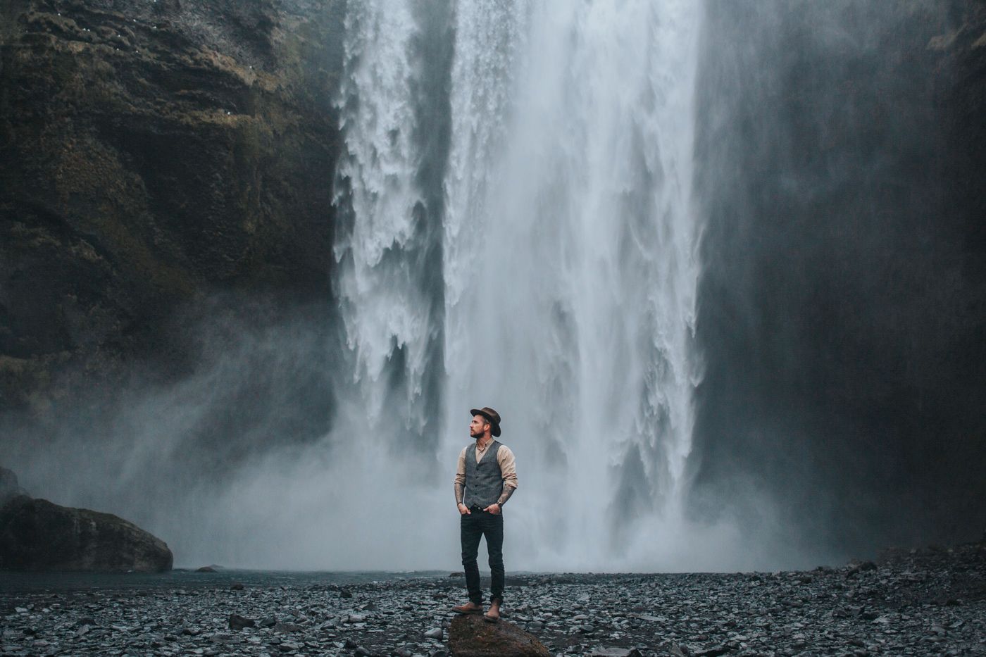 Silhouetted person stands on black beach beneath massive waterfall in Iceland.