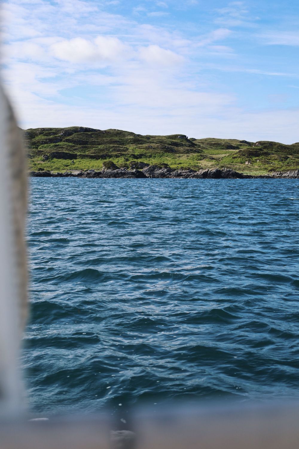 Seascape showing deep blue waters and green coastal hills in the distance.