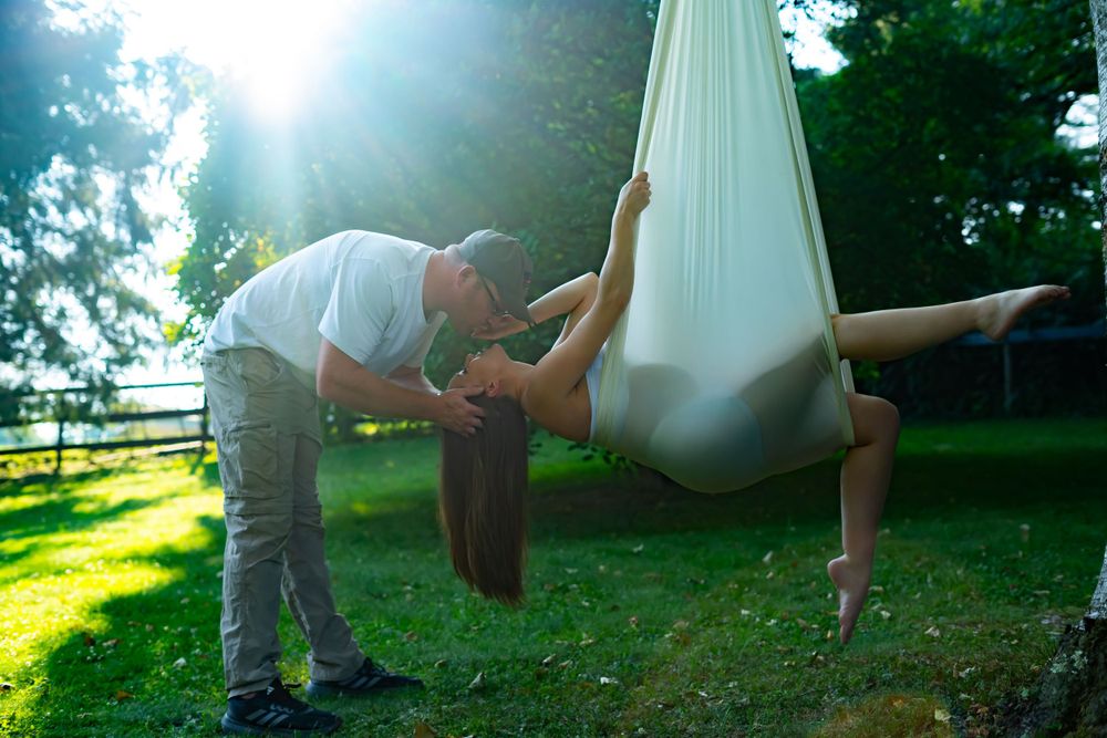 Person assists another in white aerial silk hammock during outdoor sunset session.