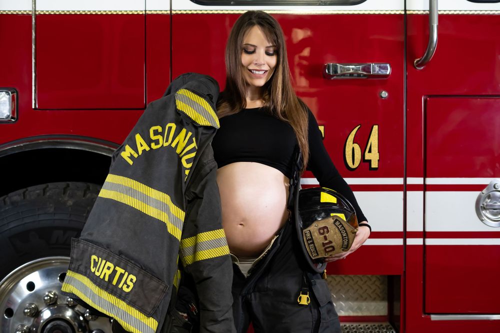 Pregnant woman wearing firefighter gear poses next to bright red fire engine number 64.