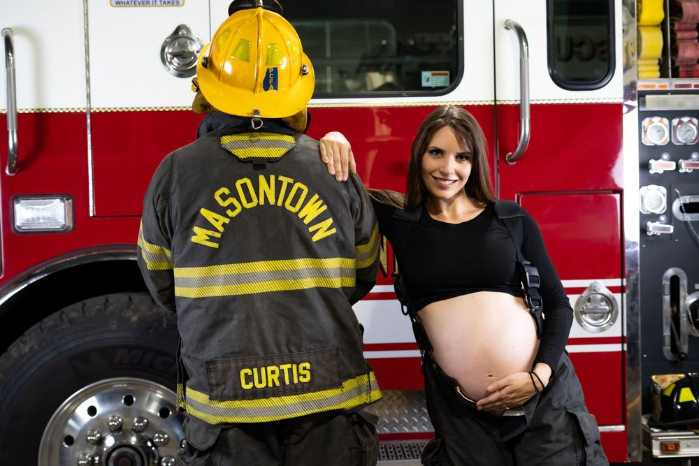 Pregnant woman stands beside firefighter in front of red fire truck.
