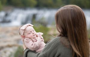 Parent in green sweater holding smiling baby in pink against waterfall scenery.