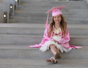 Graduate in pink cap and gown sitting and reading on concrete floor.
