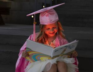 Young graduate in pink cap and gown reading a book with enthusiasm.