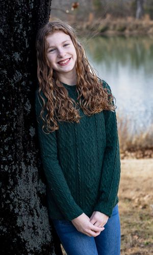 A person in a dark green cable knit sweater and jeans poses casually against a tree trunk near a lake.
